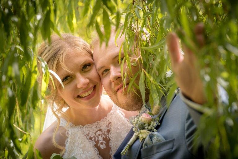Braut lacht zwischen den Zweigen einer Trauerweide – Brautpaarshooting Miriam & Pascal