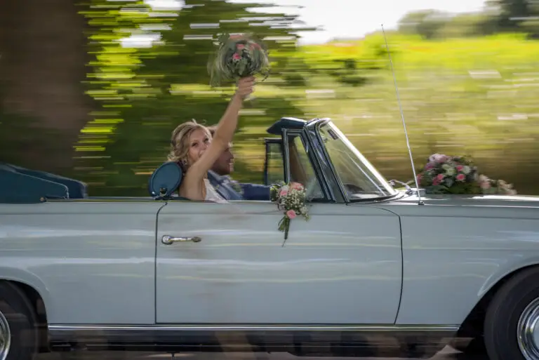 Brautpaar fährt im weißen Oldtimer-Cabrio, Braut hält den Strauß hoch – Brautpaarshooting Miriam & Pascal