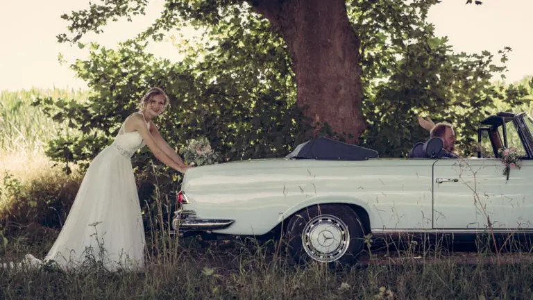 Braut lehnt an einem weißen Oldtimer-Cabrio unter einem Baum – Brautpaarshooting Miriam & Pascal
