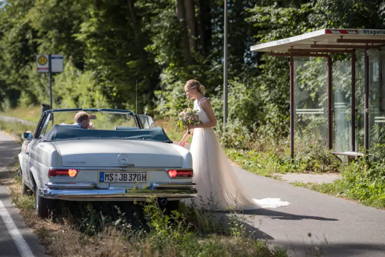Braut steht neben einem weißen Oldtimer-Cabrio am Straßenrand – Brautpaarshooting Miriam & Pascal