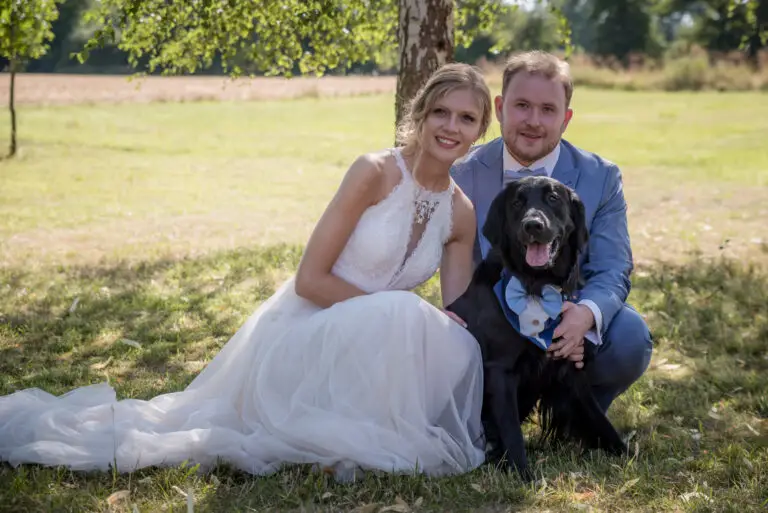 Braut im weiten Kleid sitzt mit Bräutigam und Hund auf der Wiese – Brautpaarshooting Miriam & Pascal