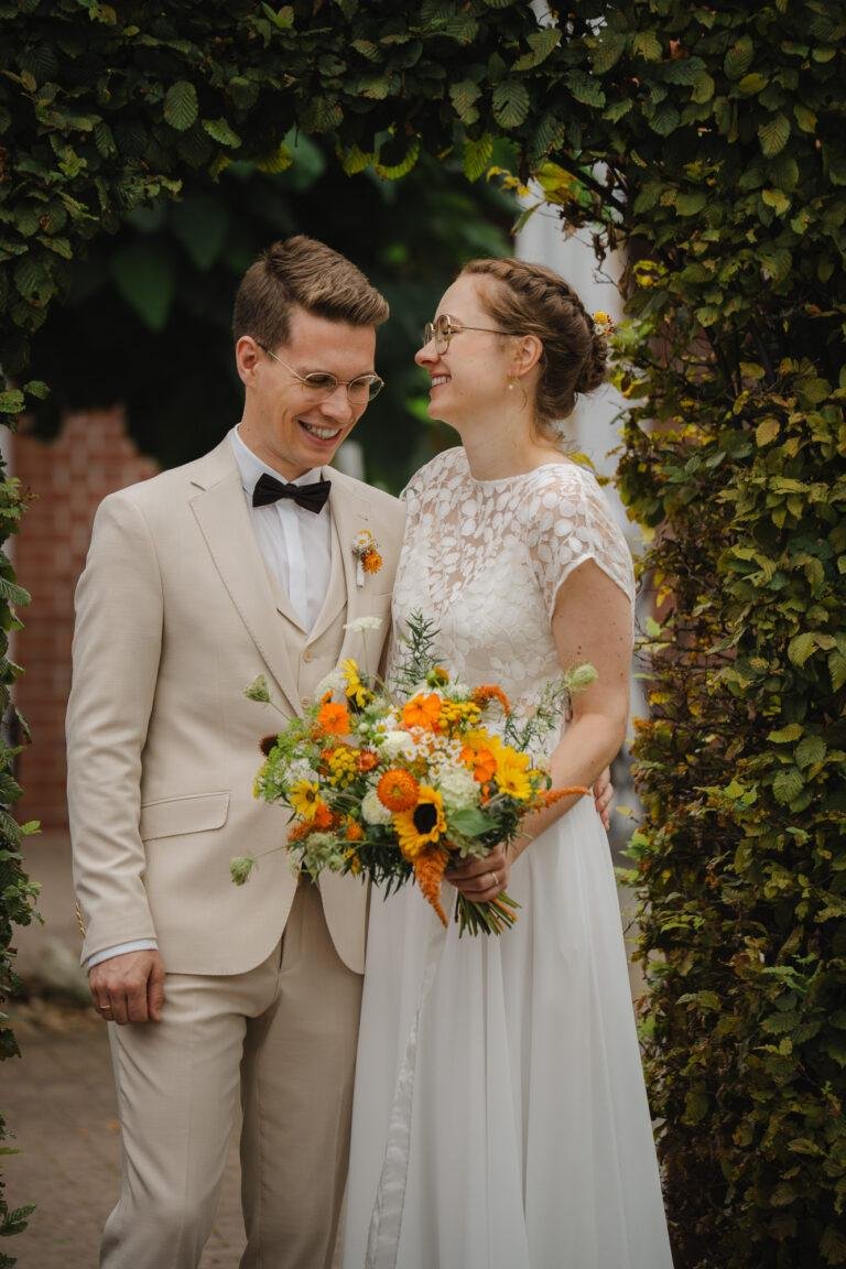 Brautpaar lacht sich mit Sonnenblumenstrauß vor einer Hecke an – Brautpaarshooting Nora & Simon