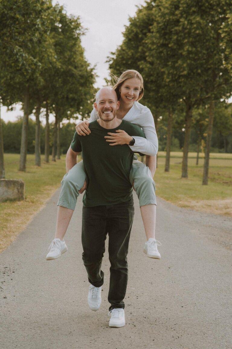 Mann trägt seine Partnerin huckepack auf einer Allee – Paarfoto von SNDR Photos in Münster