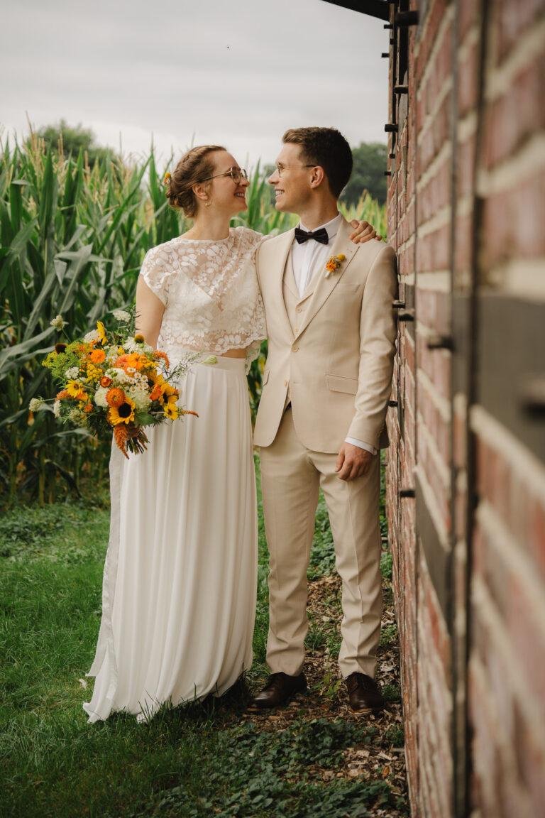 Braut mit Sonnenblumenstrauß und Bräutigam vor einem Maisfeld – Brautpaarshooting Nora & Simon