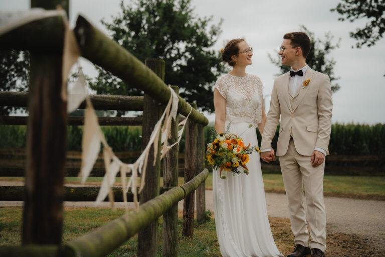 Brautpaar steht an einem Holzzaun mit Pampasgras-Dekoration – Brautpaarshooting Nora & Simon