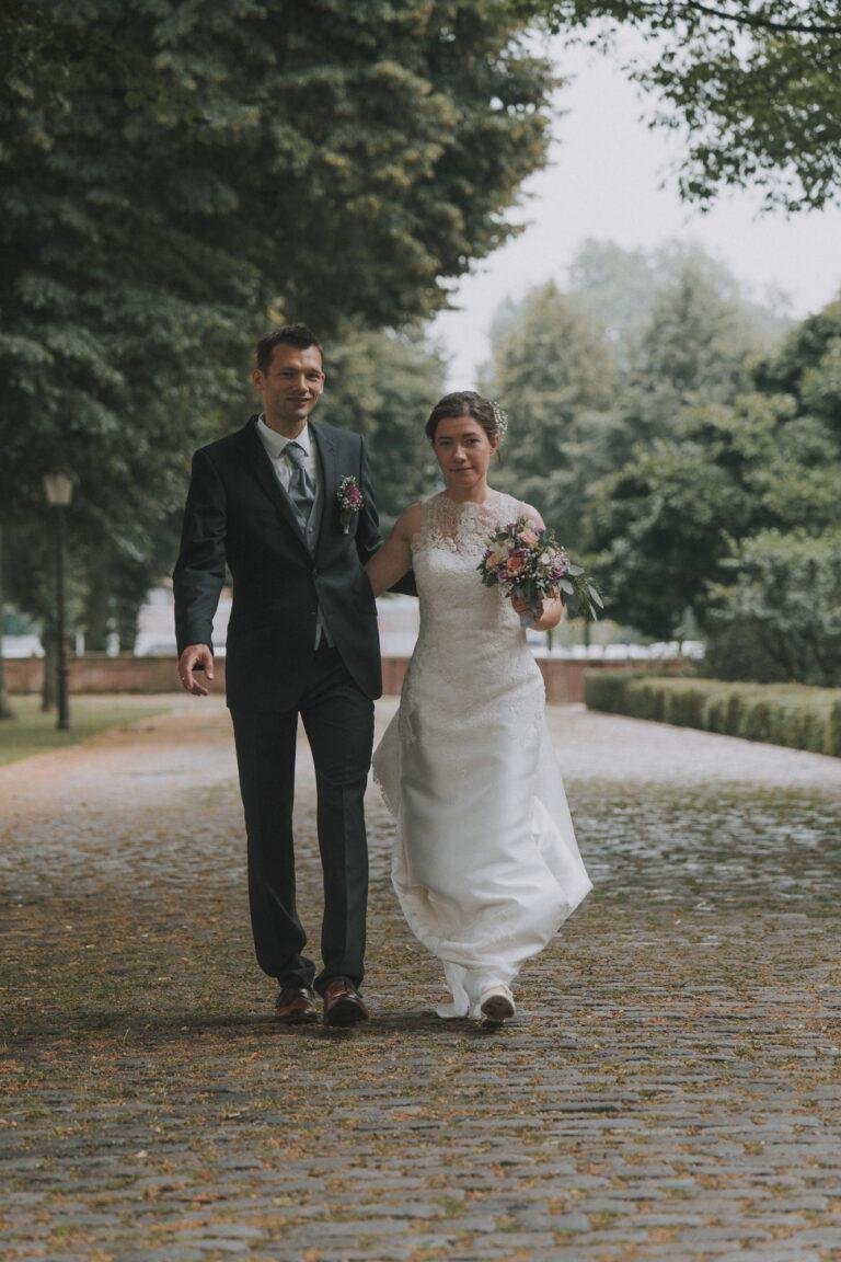 Web-16 Brautpaar geht Hand in Hand durch eine Allee – Brautpaarshooting Bernadette & André am Schloss Münster