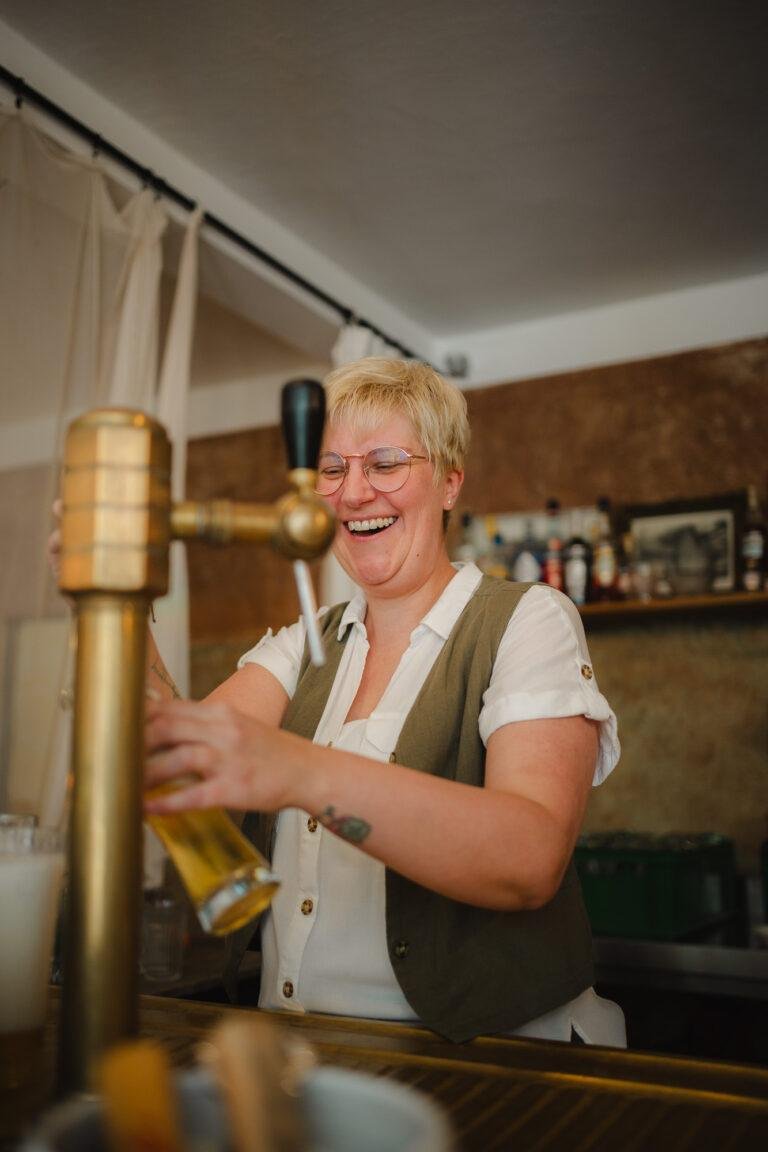 Web-17 Gastgeberin zapft lachend ein Getränk hinter der Theke – Hochzeitsfeier Renée & Patrick im Café Nobis