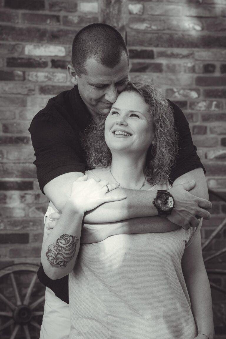 Schwarz-Weiß-Foto: Mann umarmt Frau von hinten vor einer Backsteinwand – Paarfotoshooting SNDR Photos Münster