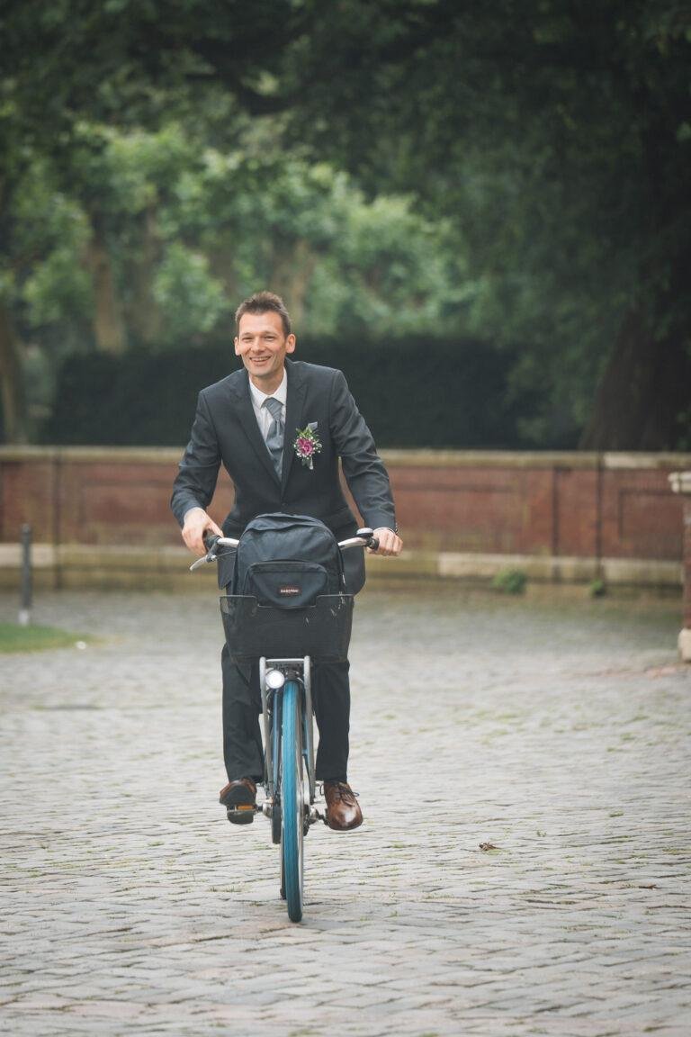 Web-3 Bräutigam fährt lachend Fahrrad über das Kopfsteinpflaster – Brautpaarshooting Bernadette & André am Schloss Münster