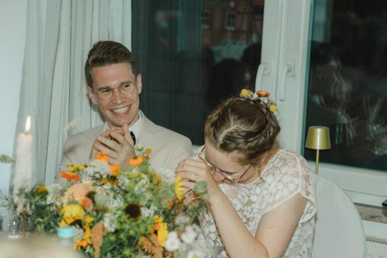 Bräutigam und Braut lachen am Hochzeitstisch mit Blumendeko – Hochzeitsfeier Nora & Simon