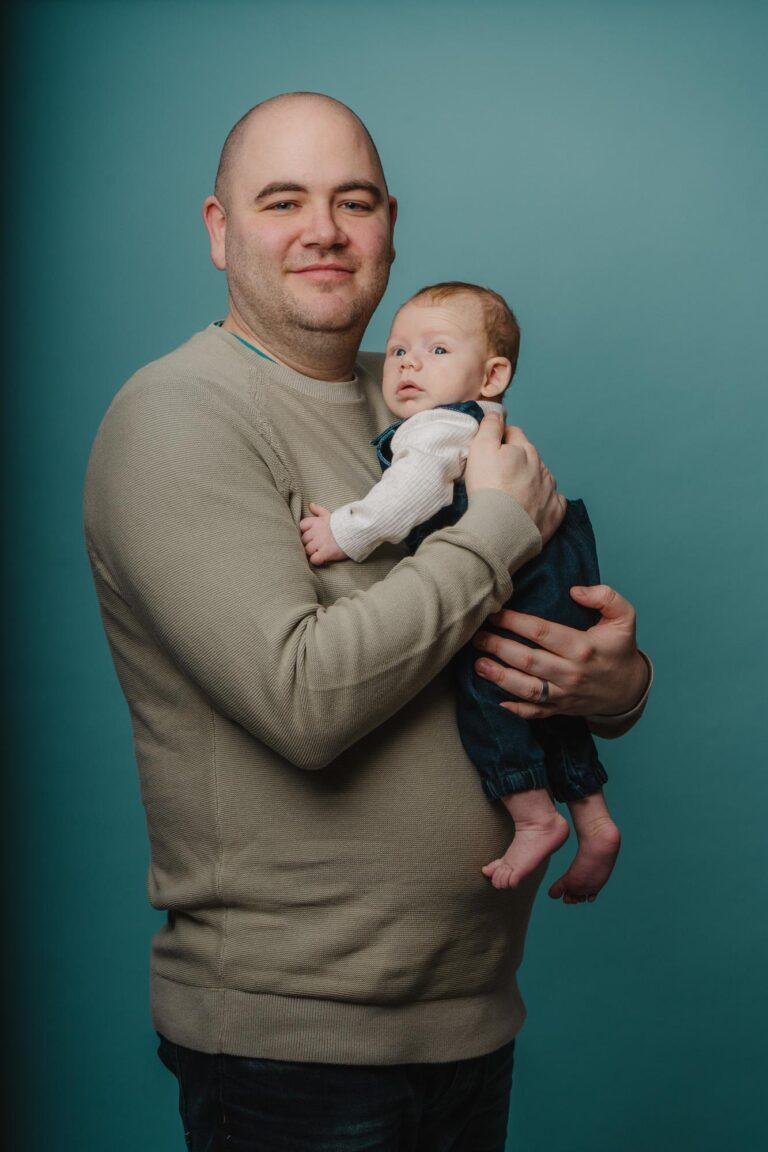 Vater hält sein Baby im Arm vor türkisem Hintergrund – Babyfotos SNDR Photos Münster