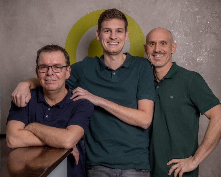 Drei Männer im Team posieren vor einem Logo an der Wand – Business-Fotografie SNDR Photos Münster